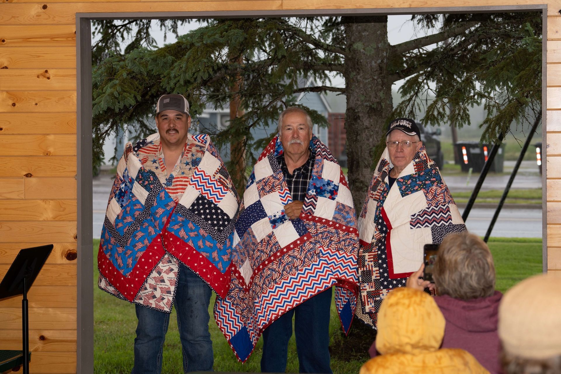 Three singers in quilt-pattern vests perform on an outdoor stage in front of an audience.