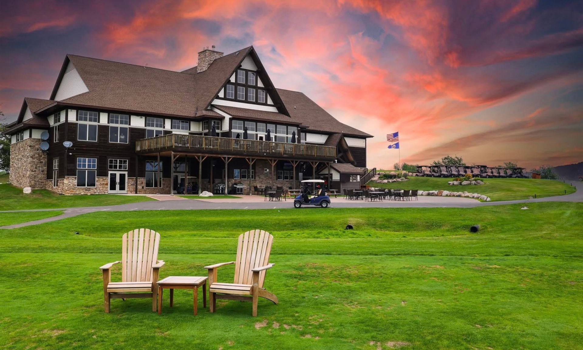 Two wooden chairs on a green lawn before a large lodge at sunset, with dramatic pink-orange sky.