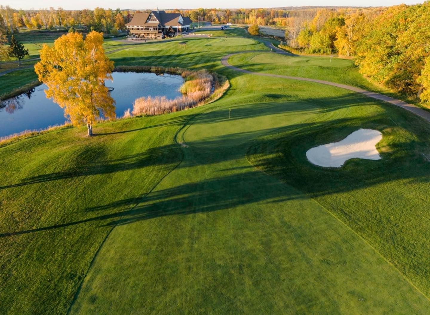Golf course with green fairways, sand bunker, pond, and autumn trees at sunset