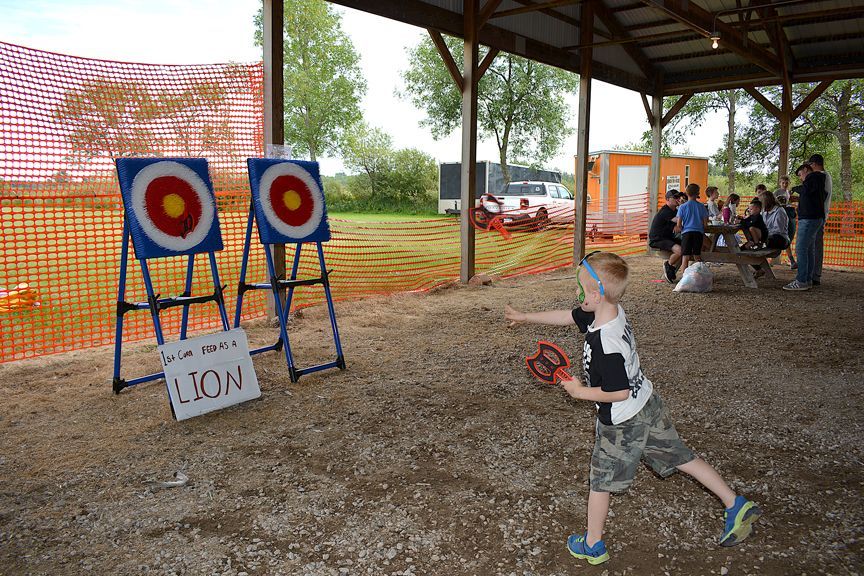 Child practicing archery at outdoor range with targets and onlookers under a pavilion