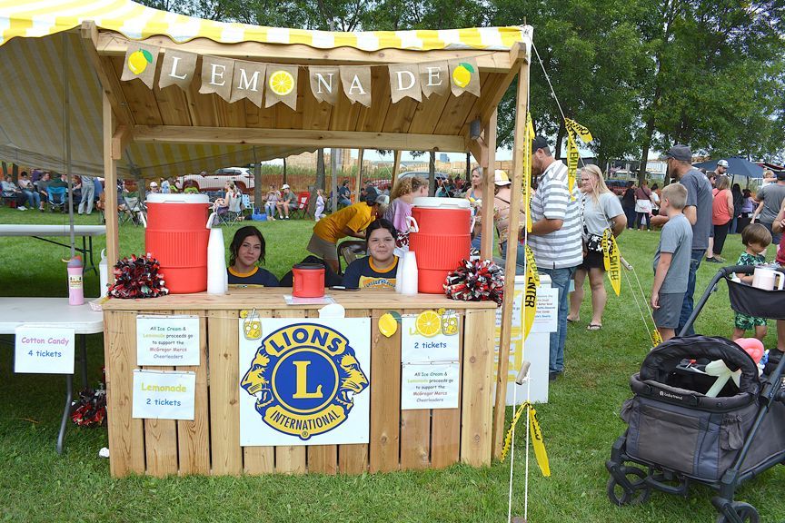 Lemonade stand with children serving drinks at a park fair, decorated with Lions Club signs and red coolers