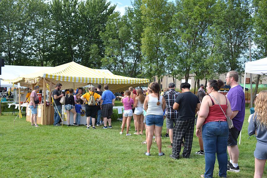 People standing in line at an outdoor fair under striped tents on a grassy field