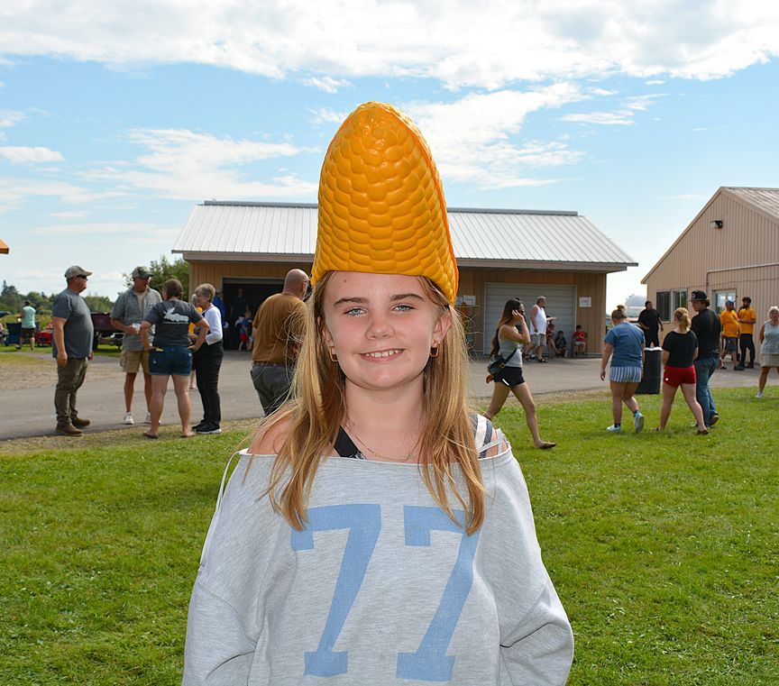 Person wearing a tall orange corn hat stands in a grassy field at an outdoor event.