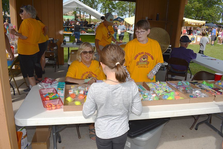 People selling colorful items at a fair booth while a visitor browses the table