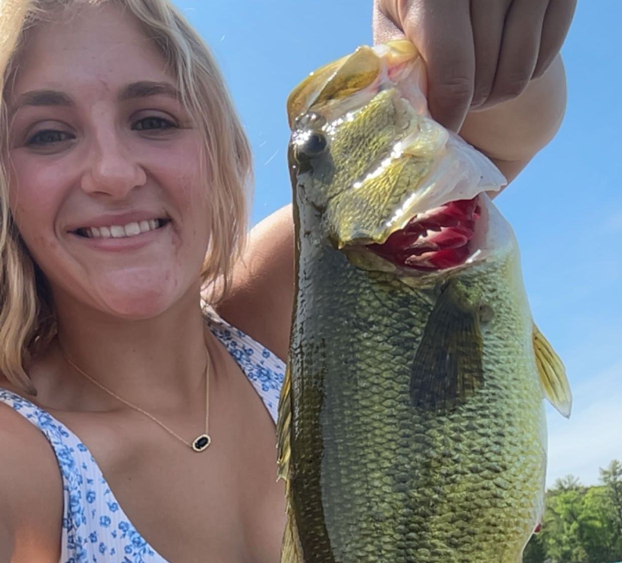 Smiling woman holding a large bass fish outdoors under bright sunlight