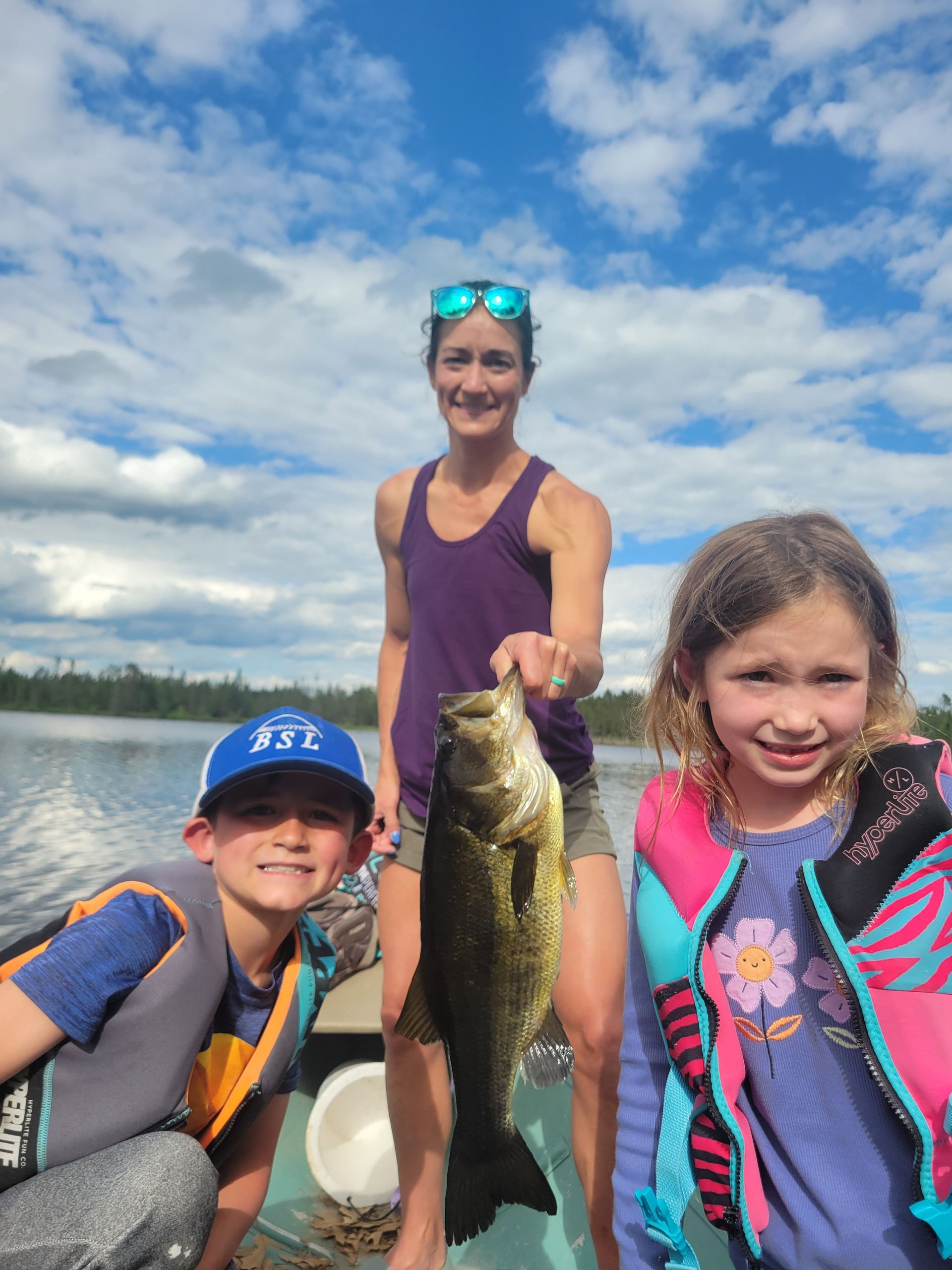 Three people on a boat holding up a large fish on a lake under a cloudy sky.
