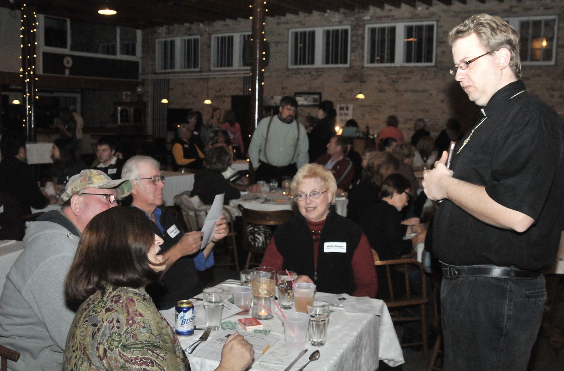Man speaking to seated audience at a crowded indoor event with round tables and drinks
