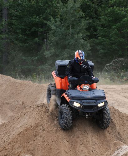 Person riding an orange ATV over a dirt mound in a wooded off-road area