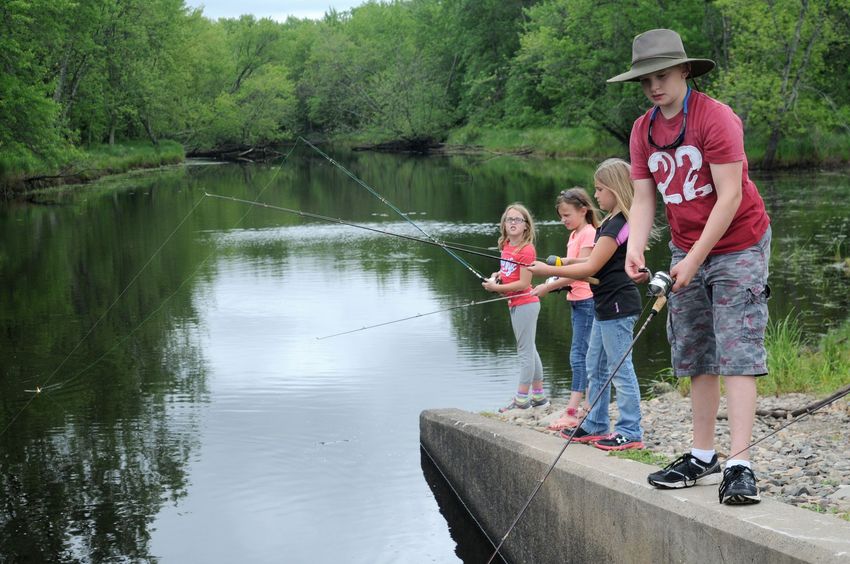 Three people fishing from a concrete dock beside a calm, tree-lined river