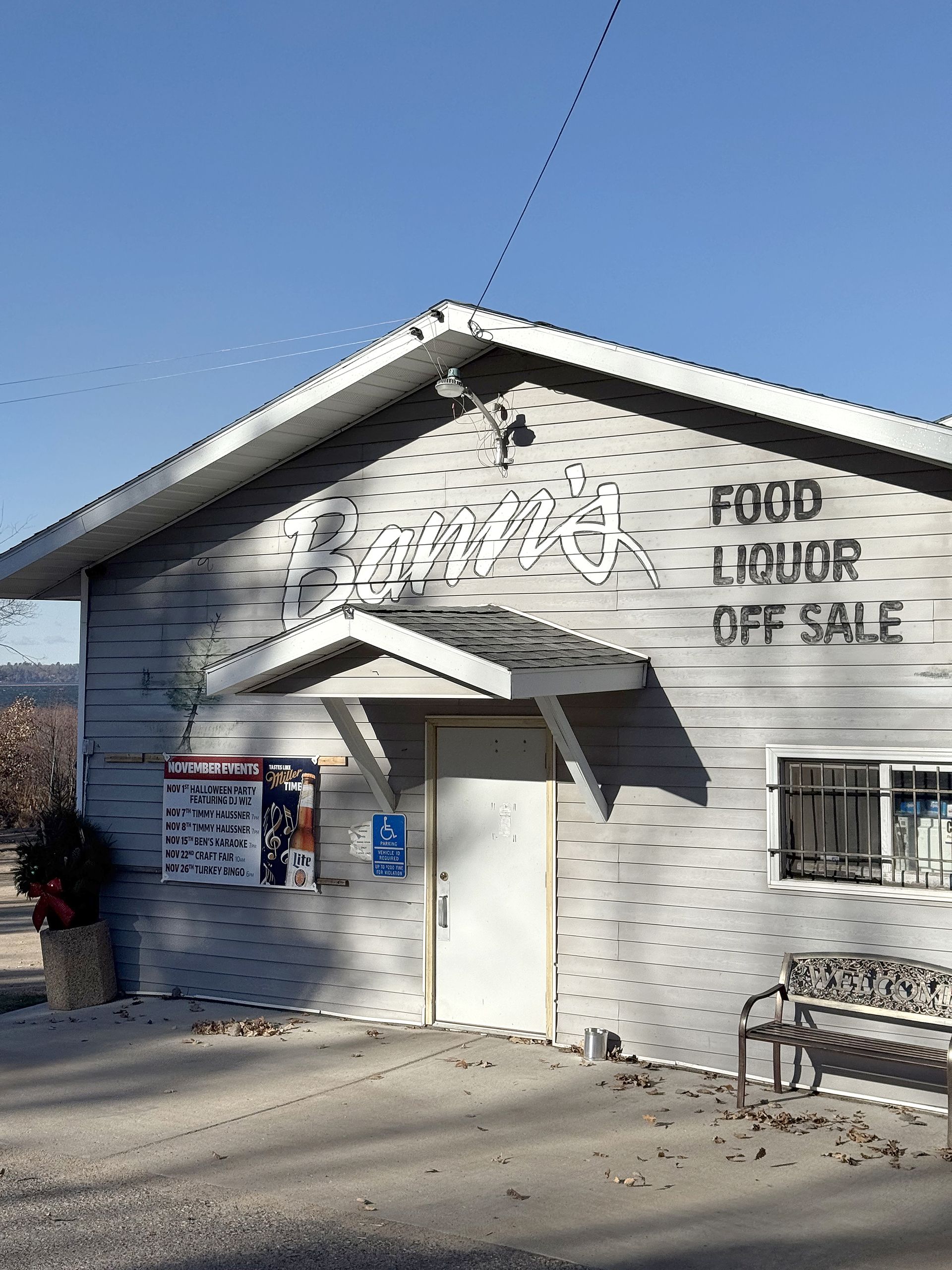 Small white building with sign, front door, windows, and bench on a sunny street corner.