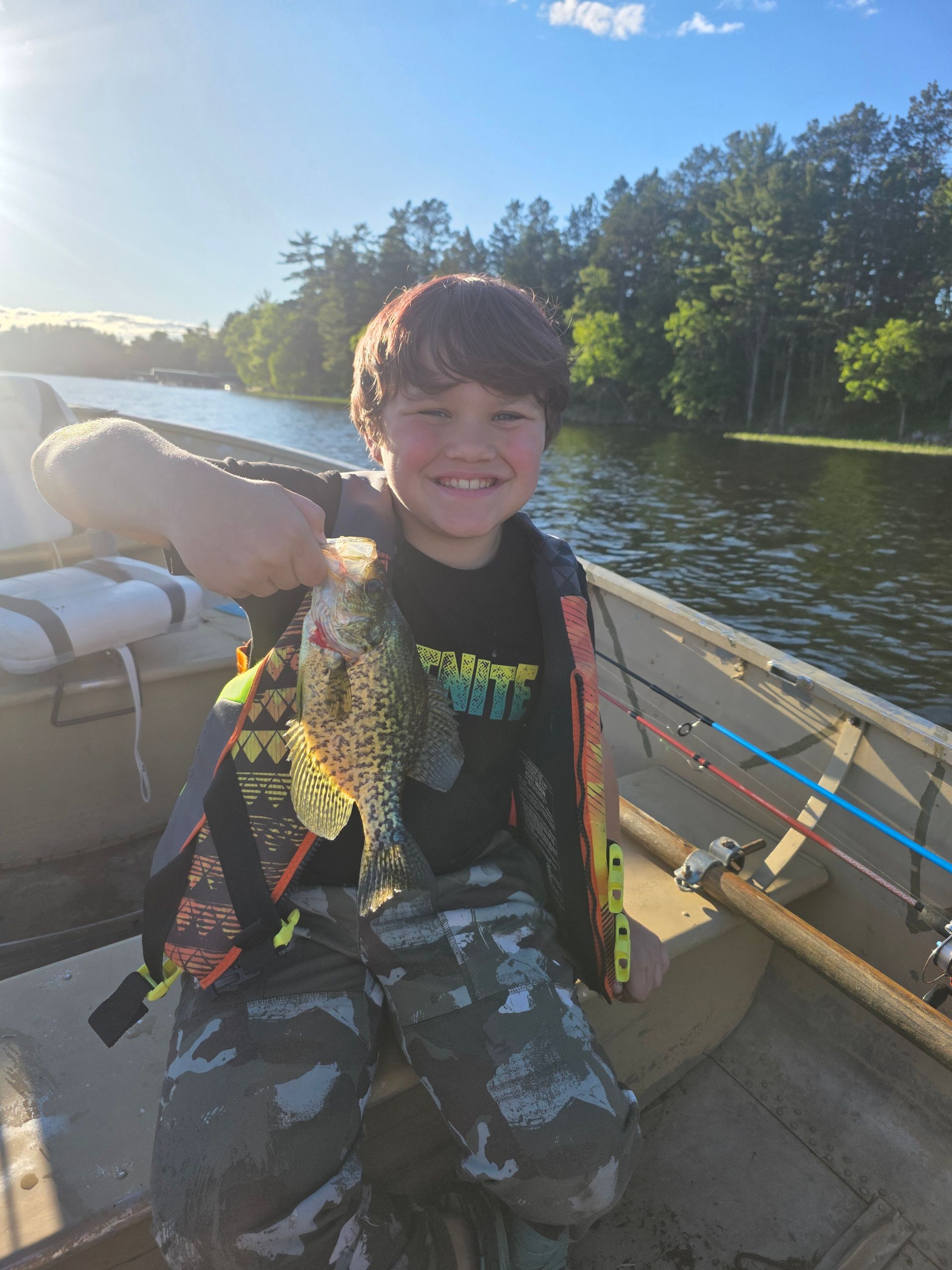 Smiling child in a boat holding a fish on a sunny lake, with fishing rods and trees nearby