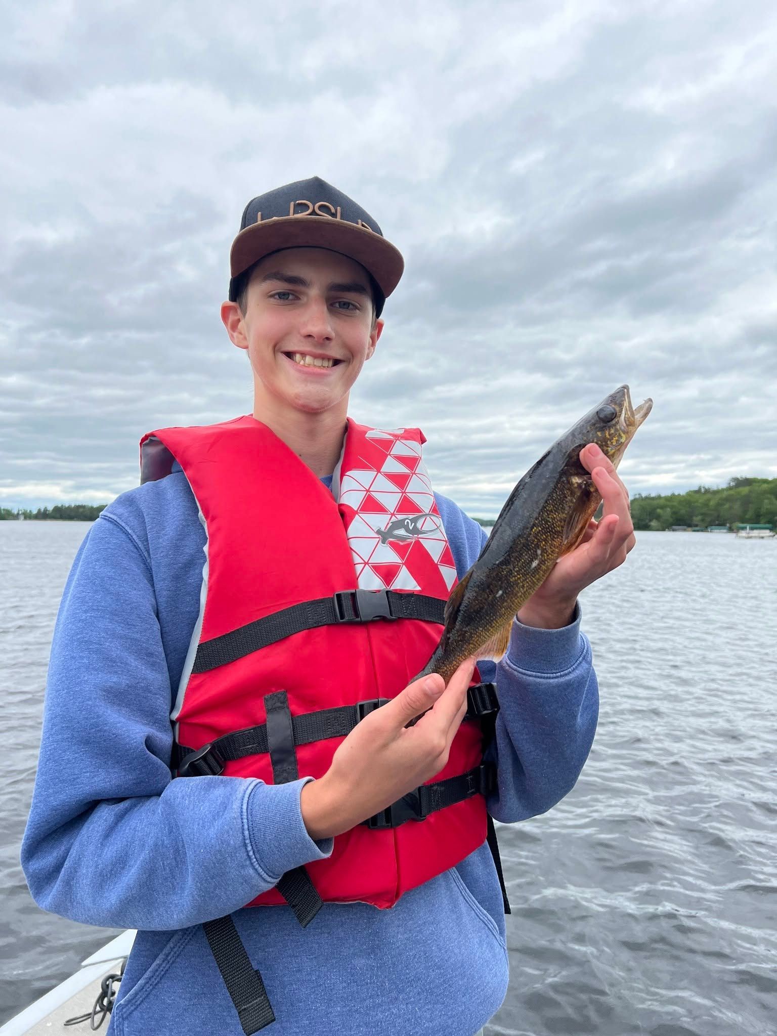 Person in a red life vest holding a fish on a boat on a cloudy lake