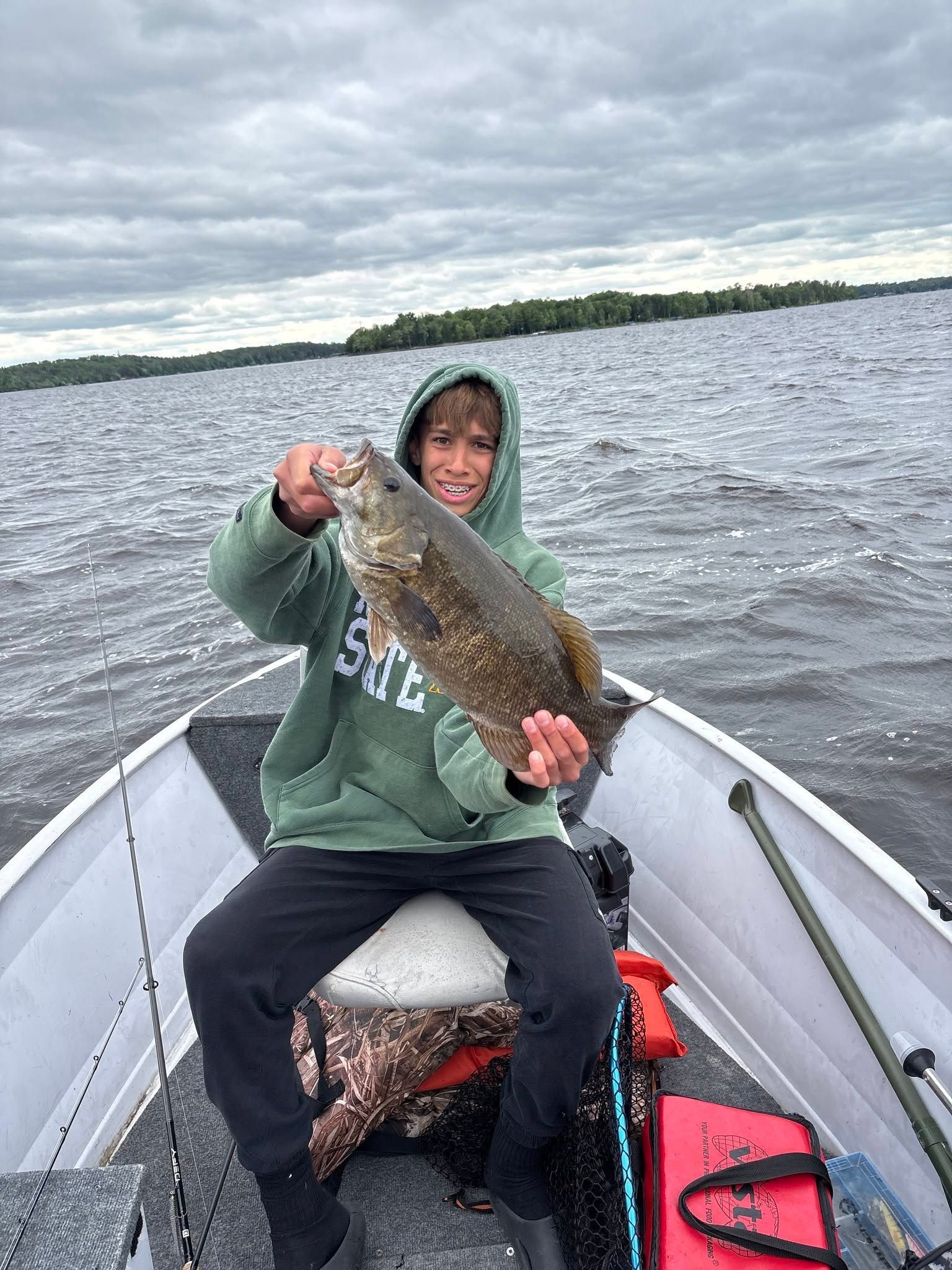 Person in a hoodie on a boat holding up a large fish on a cloudy lake.