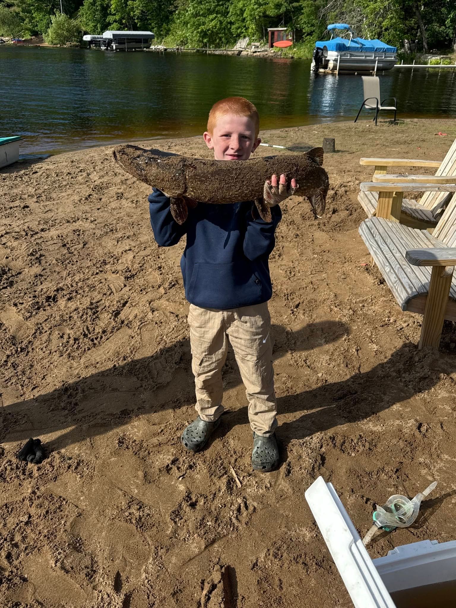 Child holding a large fish on a sandy lakeshore with boats in the background