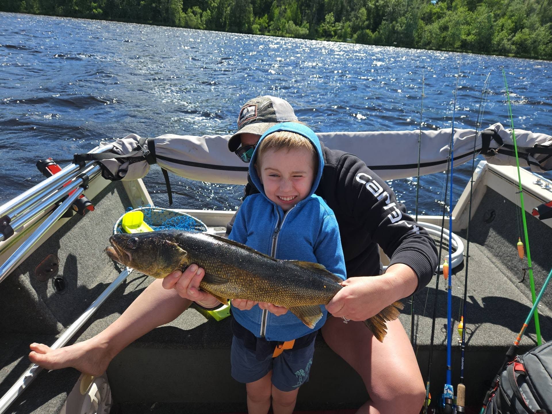 Child in a blue hoodie holding a large fish on a boat with an adult, on a lake.