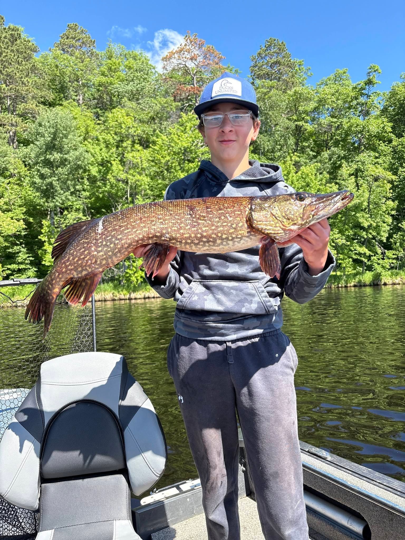 Person on a boat holding a large fish on a sunny lake with trees in the background