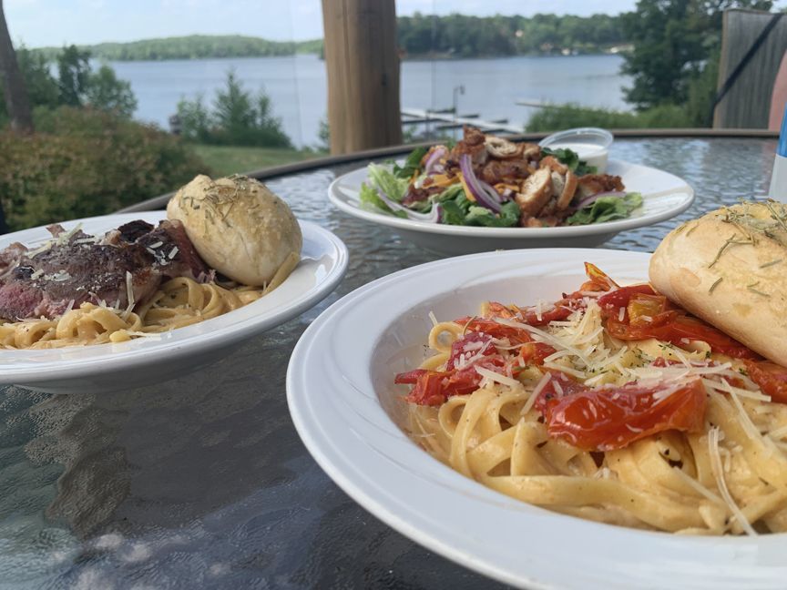 Three plates of pasta with bread on a table overlooking a lake and trees