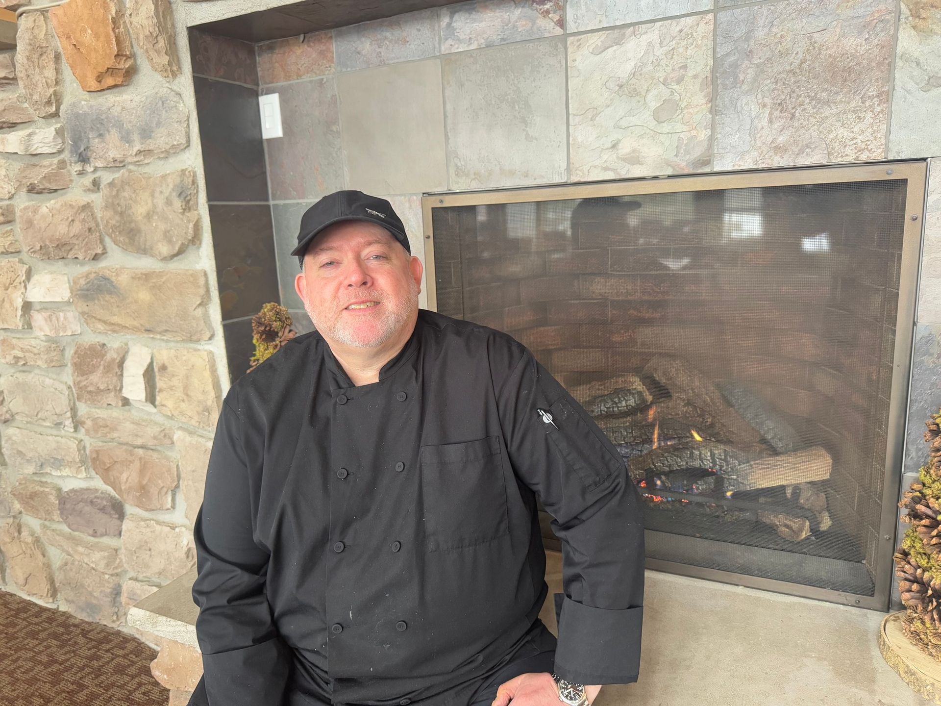Person in black chef coat and cap sitting beside a stone fireplace with a fire burning.