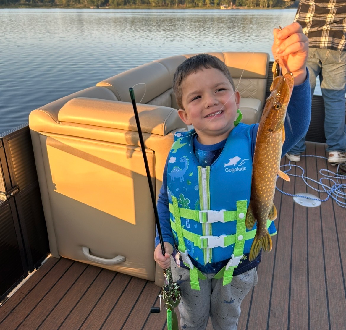Smiling child holding a fish on a boat deck at sunset, wearing a blue life jacket