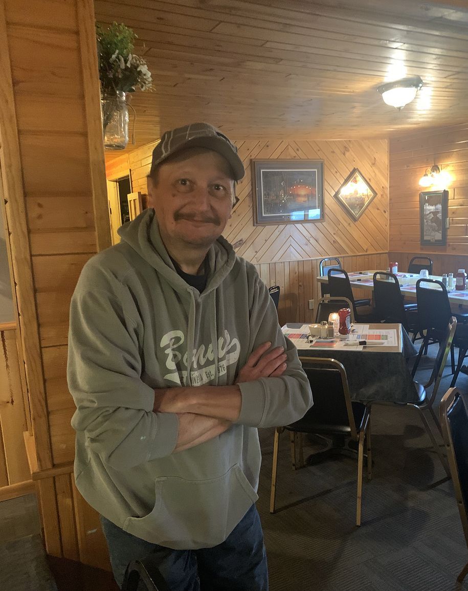 Man in gray hoodie and cap stands with arms crossed in a cozy wood-paneled restaurant.