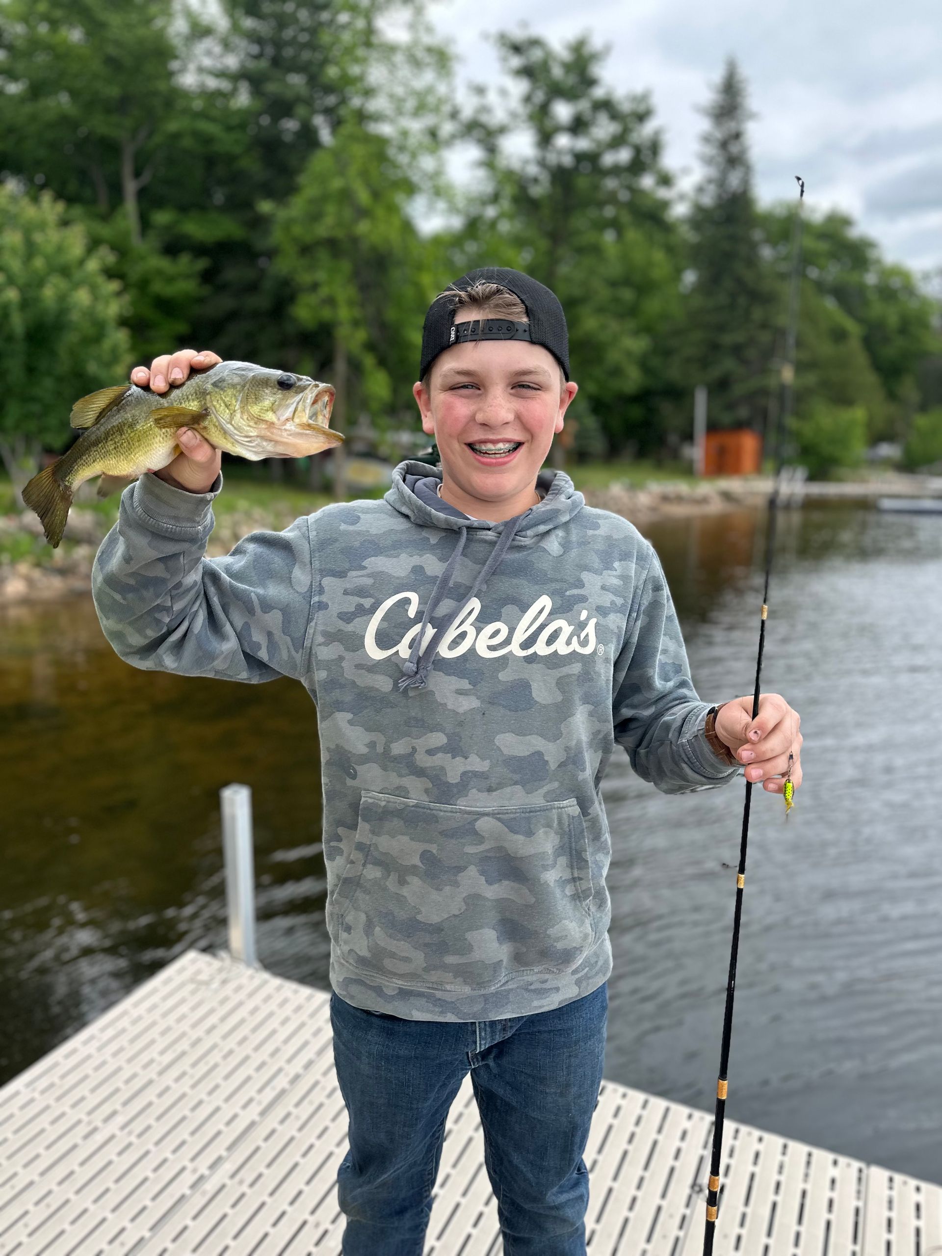 Person on a dock holding up a fish and a fishing rod beside a lake, wearing a gray Cabela’s hoodie.
