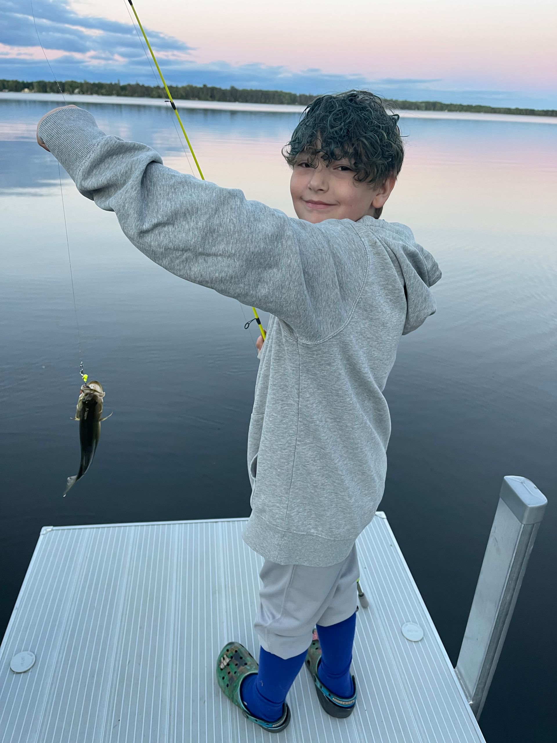 Child on a dock holding a fishing line with a fish, standing by calm water at sunset.