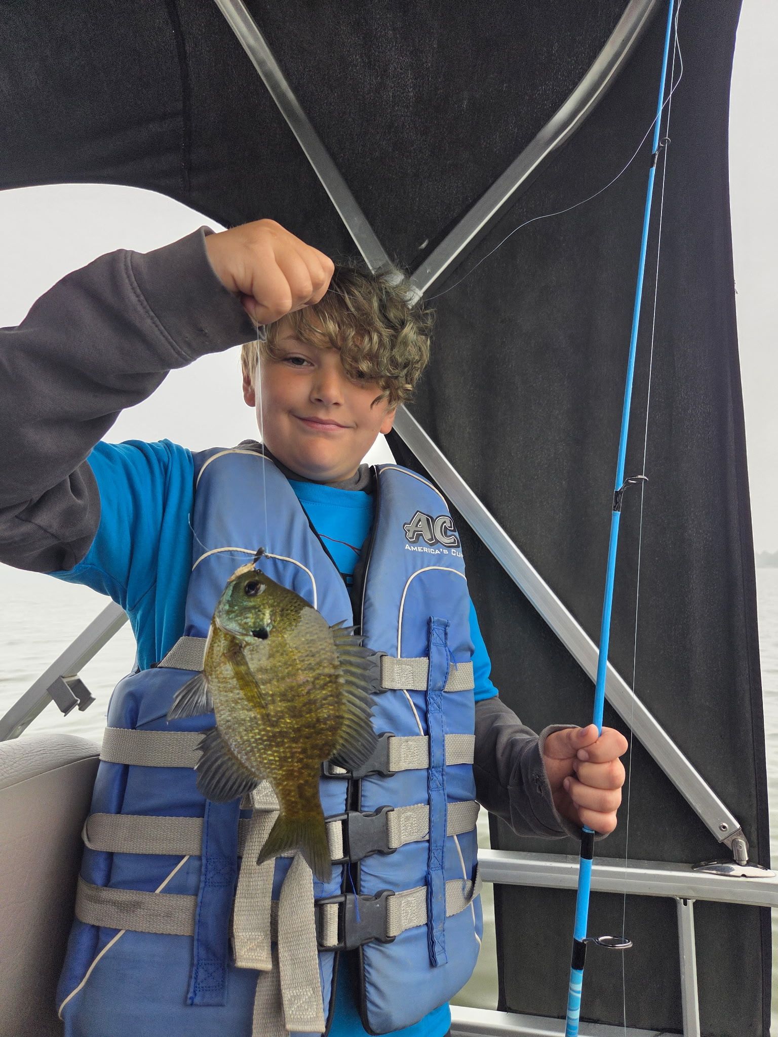 Child in a blue life vest holds up a fish on a fishing rod inside a boat or tent shelter.