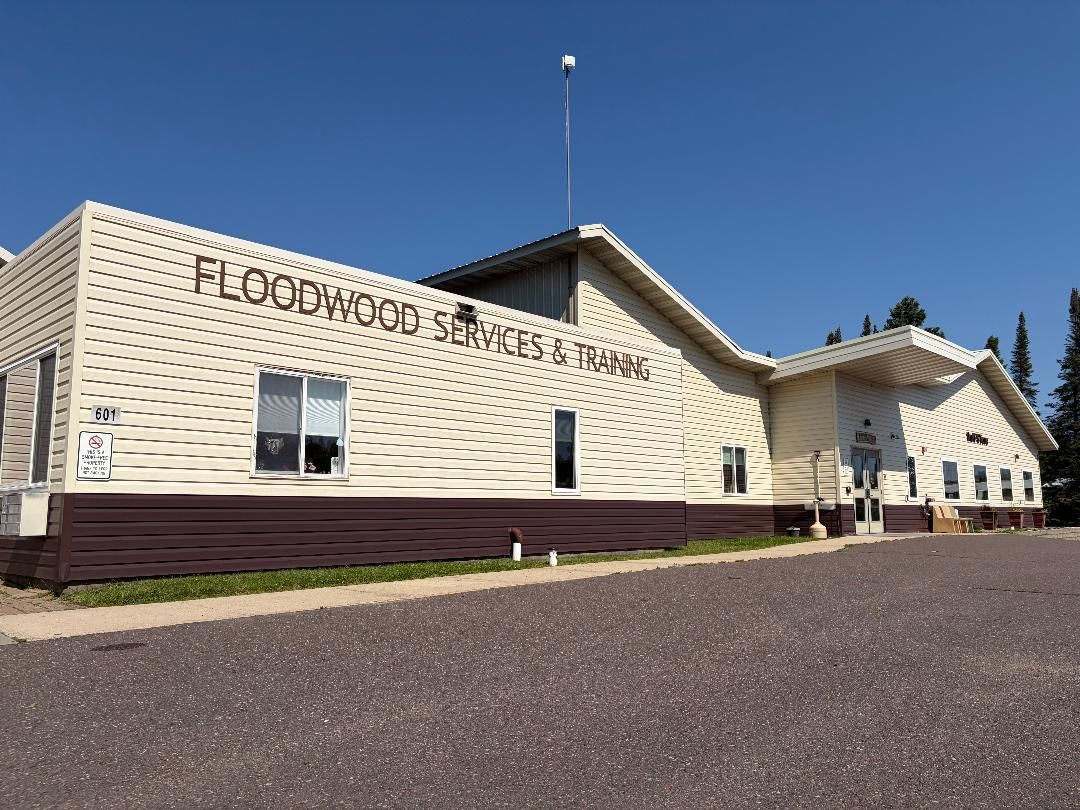 Cream-colored building with sign reading “FLOODWOOD”, viewed from a gravel lot under a blue sky.