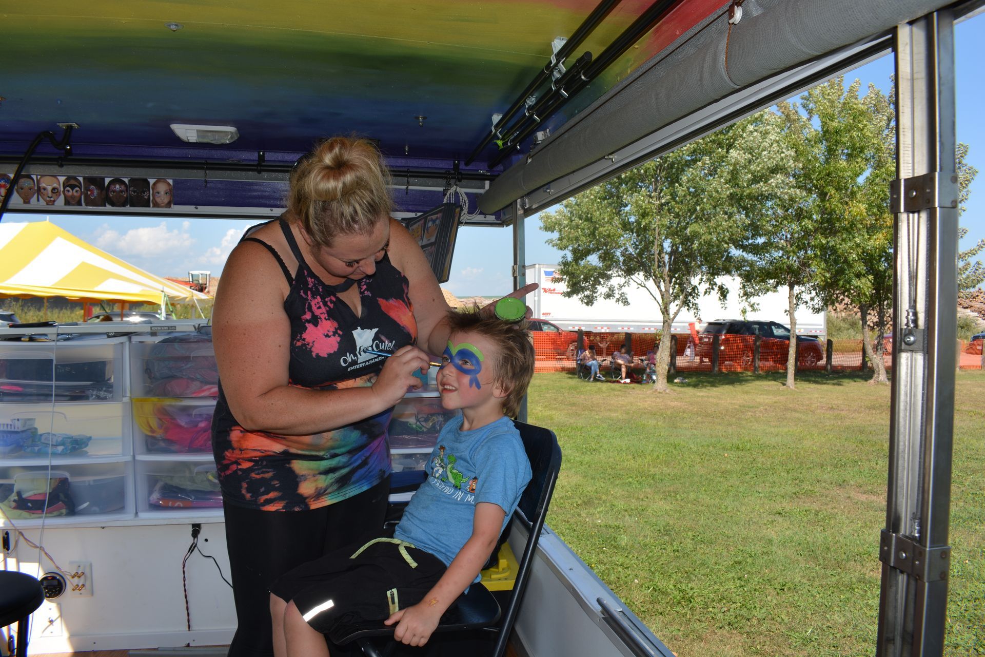 Woman braids a child’s hair inside a colorful outdoor booth.