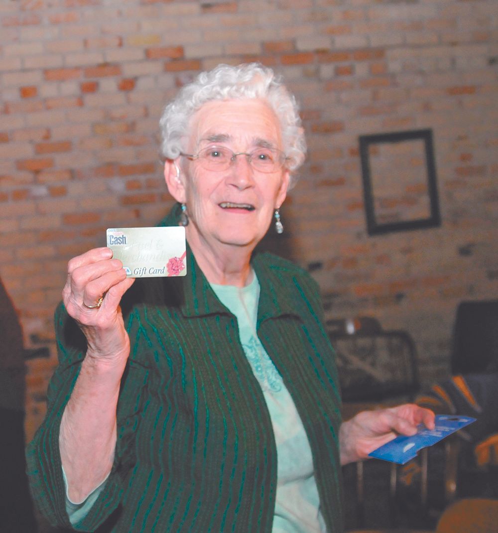 Elderly woman in a green sweater holding up a card in a brick-walled room