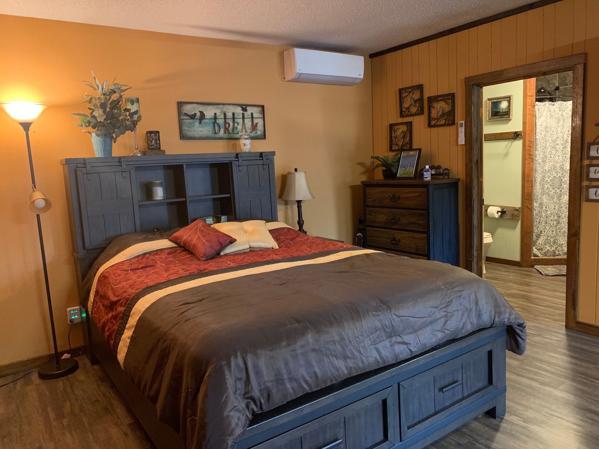 Bedroom with a blue bed, brown walls, a dresser, floor lamp, and open doorway to a bathroom.