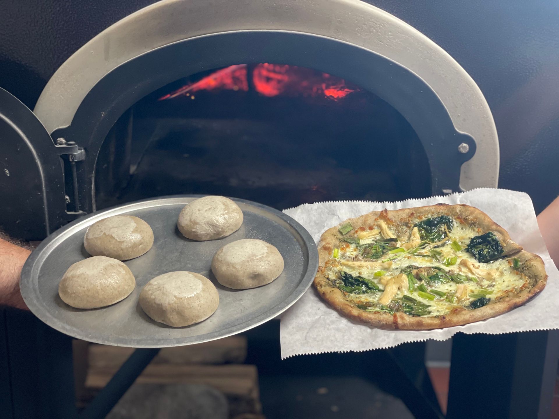 Pizza baking in a wood-fired oven beside a tray of five dough balls
