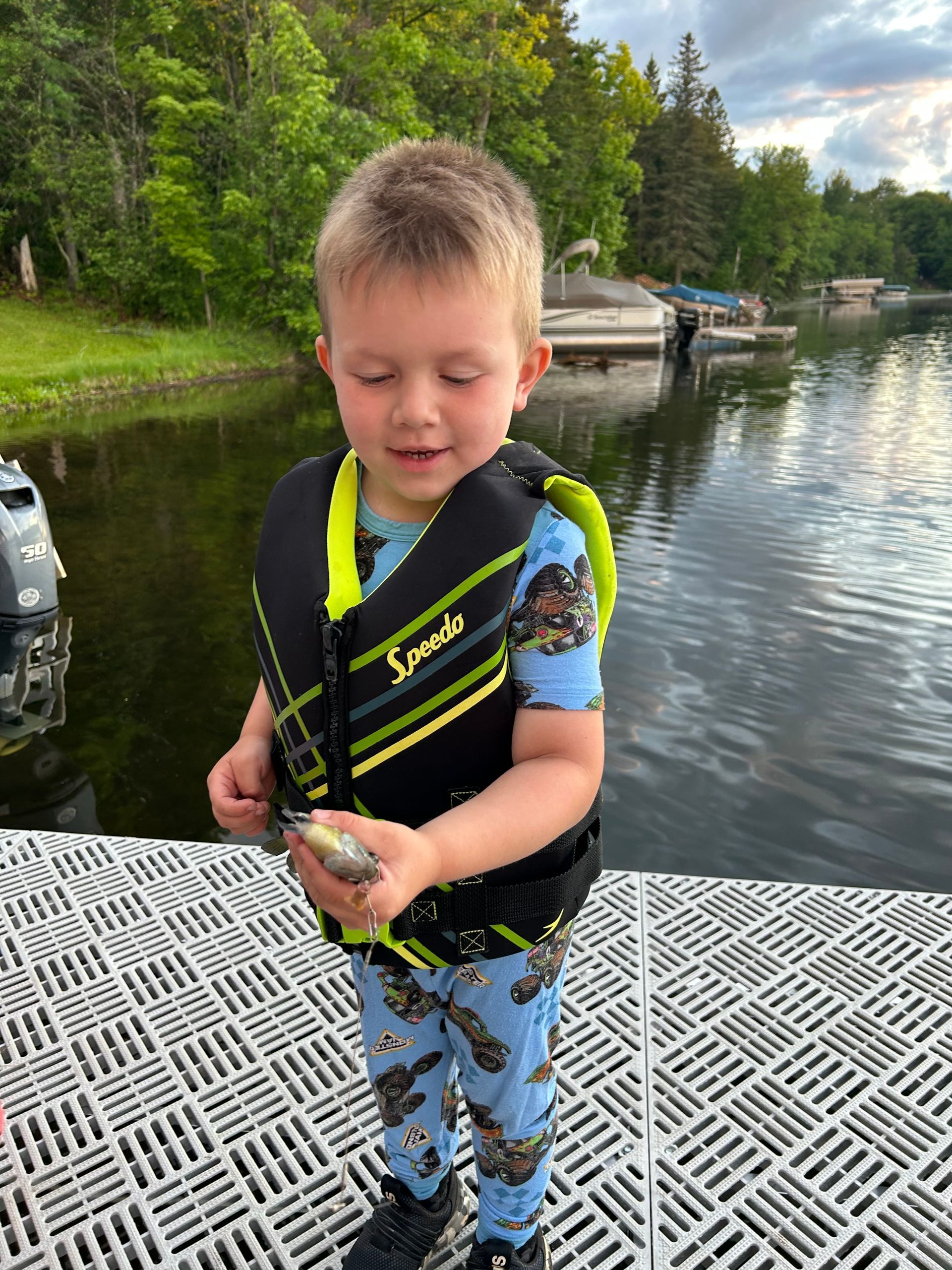 Young child in a life vest and swim shirt holding a fish by a lakeside dock