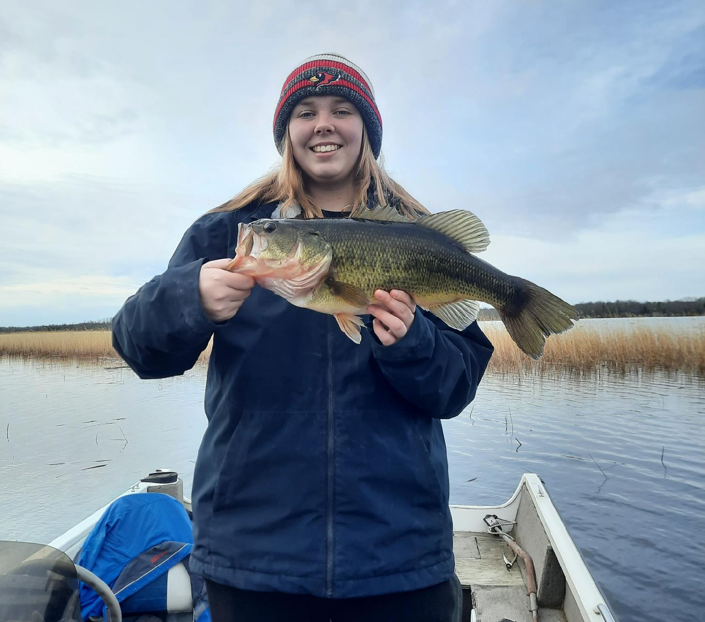 Person on a boat holding a large fish, smiling on a calm lake under a cloudy sky