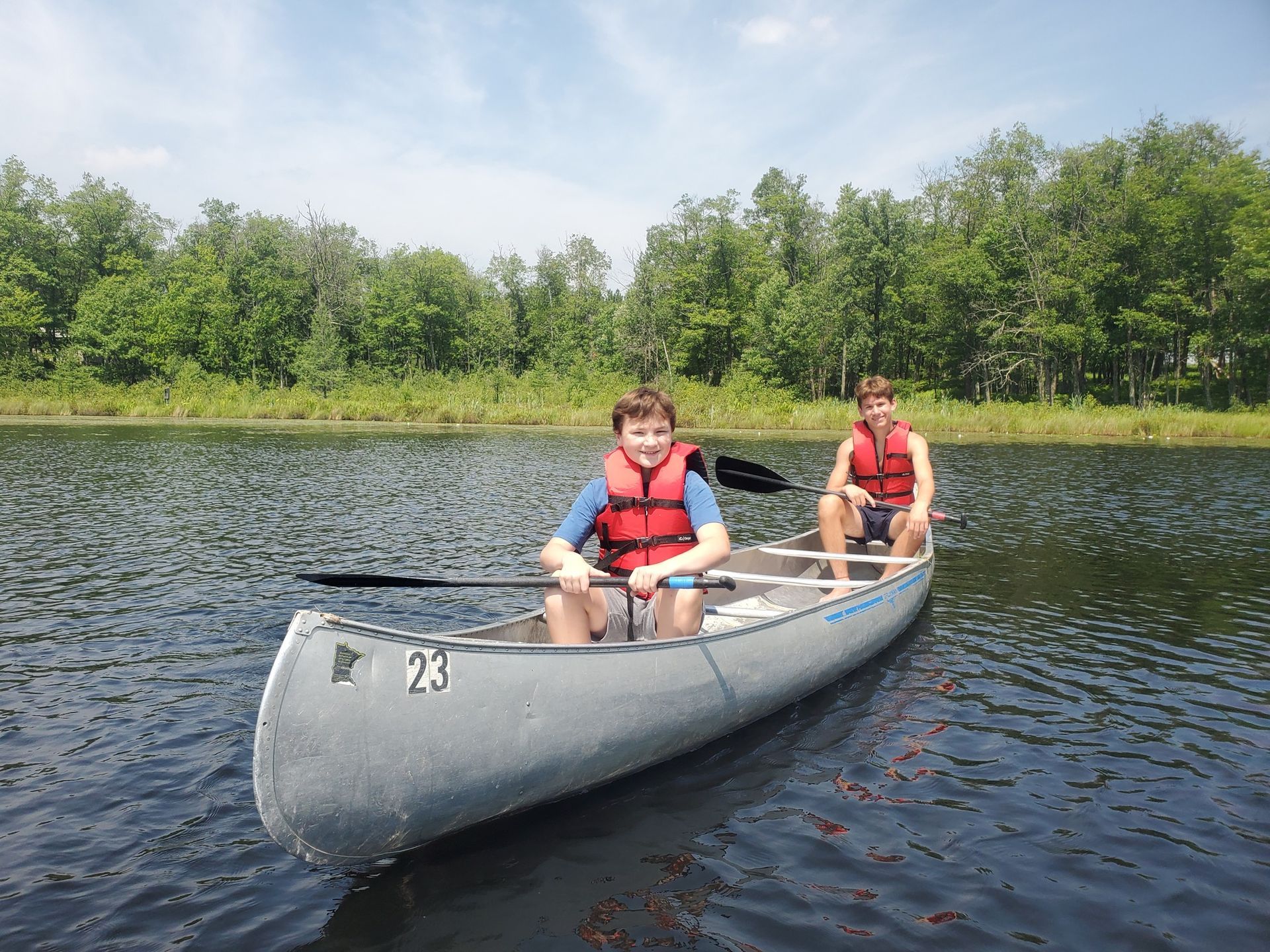 Two people in a canoe on a lake, wearing red life jackets, with trees along the shore.