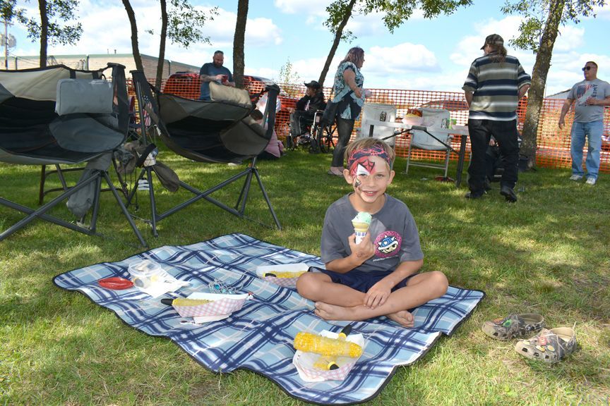 Child sitting on a picnic blanket in a park, eating ice cream with a grill and people in the background