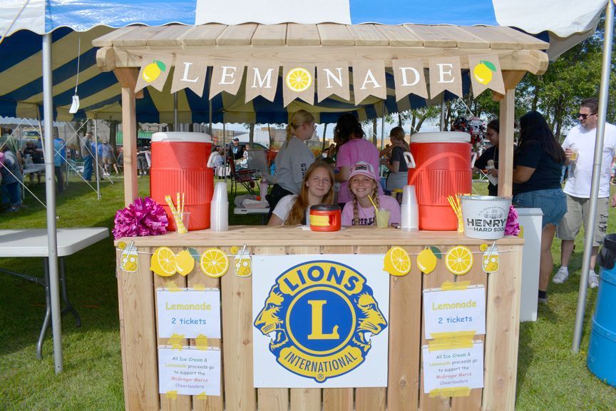 Lemonade stand with children, red drink coolers, and Lions Club sign at an outdoor fair booth