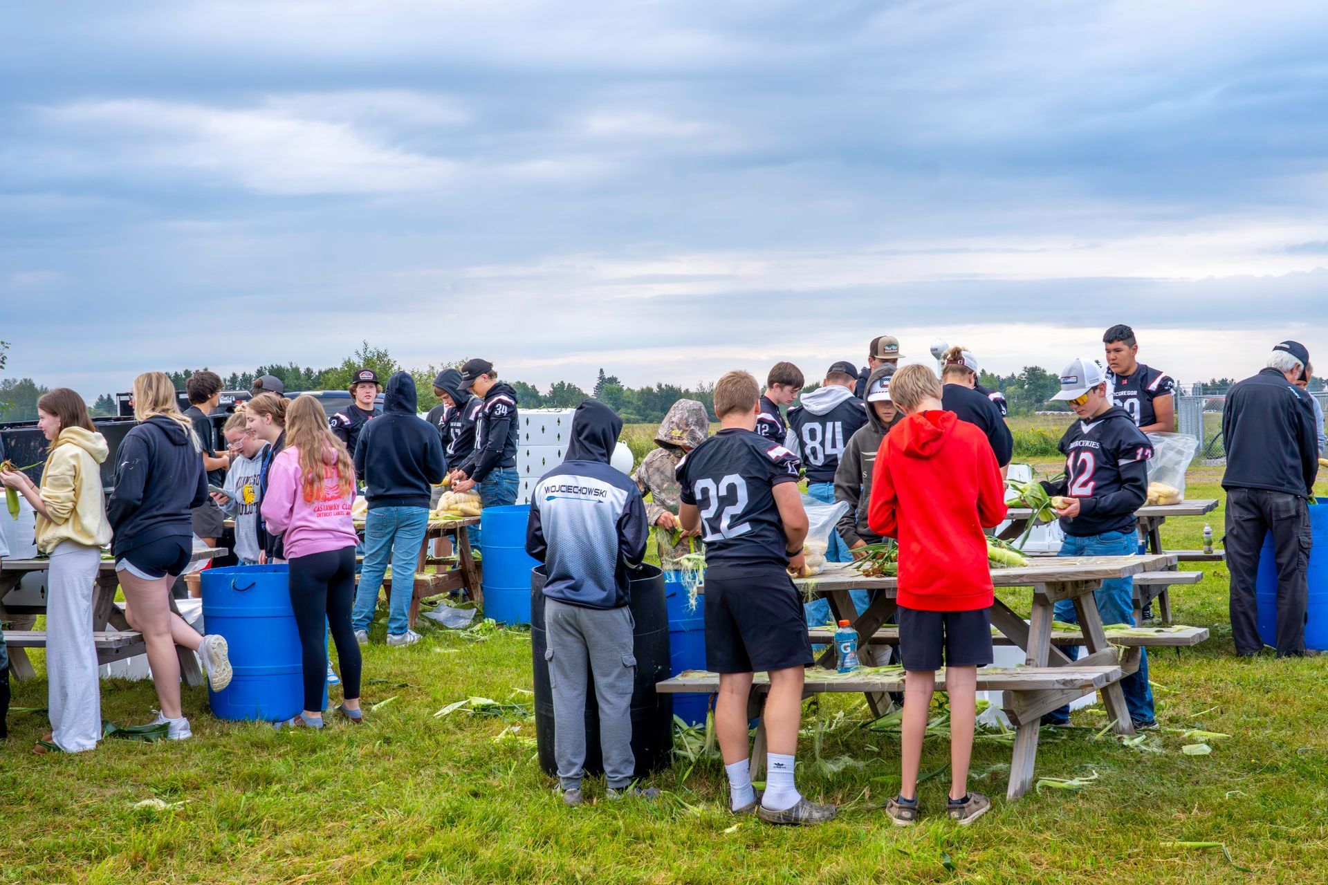 People gathered outdoors near picnic tables under a cloudy sky, some wearing sports jerseys.