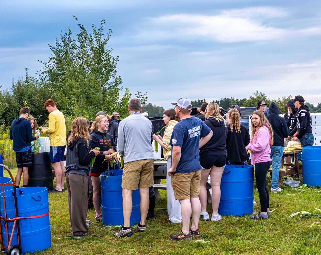 People socializing outdoors beside blue barrels on a grassy field under a cloudy sky.
