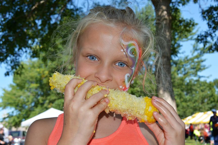 Person with face paint eating a corn cob outdoors at a fair