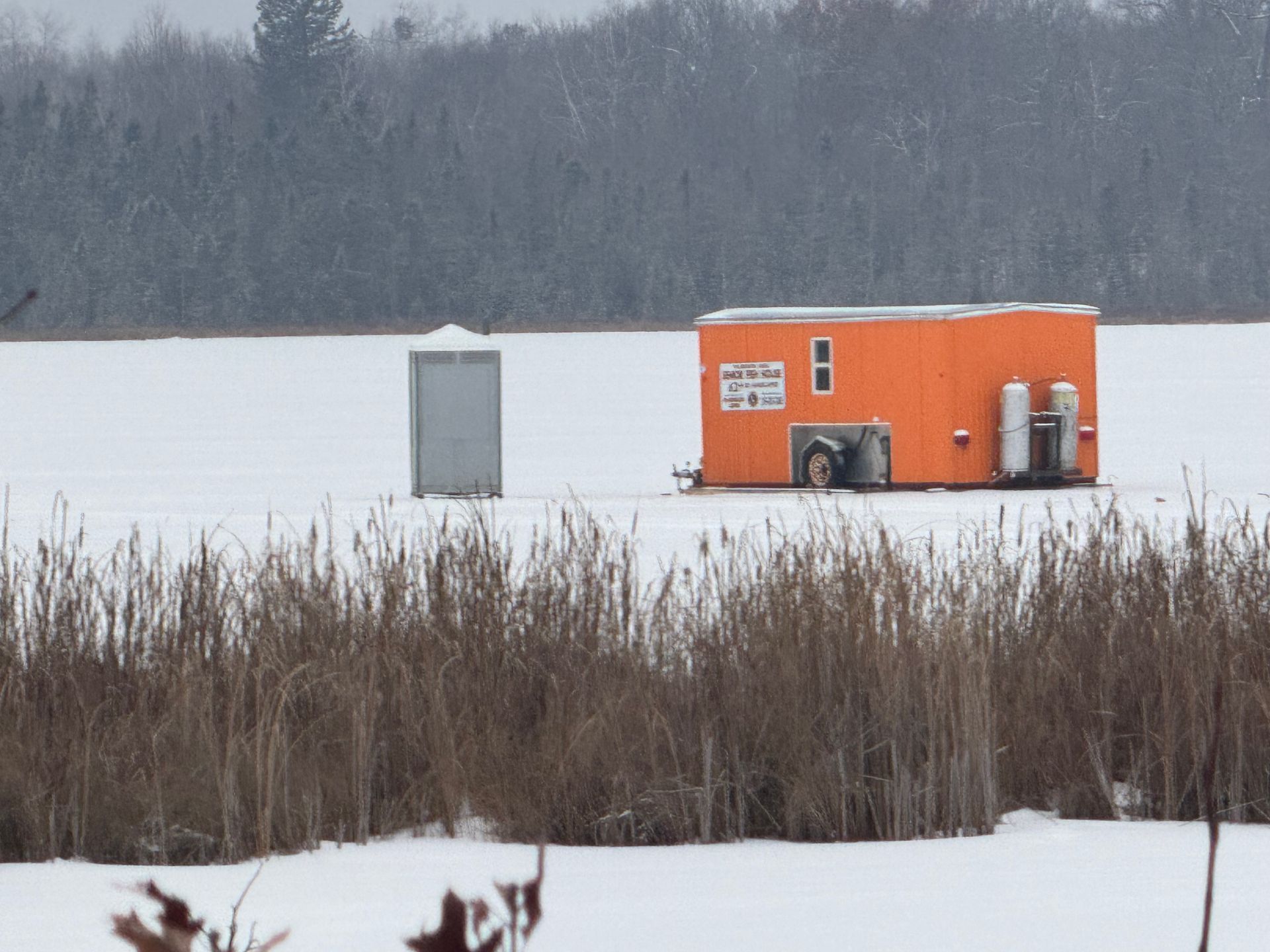 Orange ice-fishing hut on a frozen lake beside reeds and a gray shelter, with snowy woods in the background