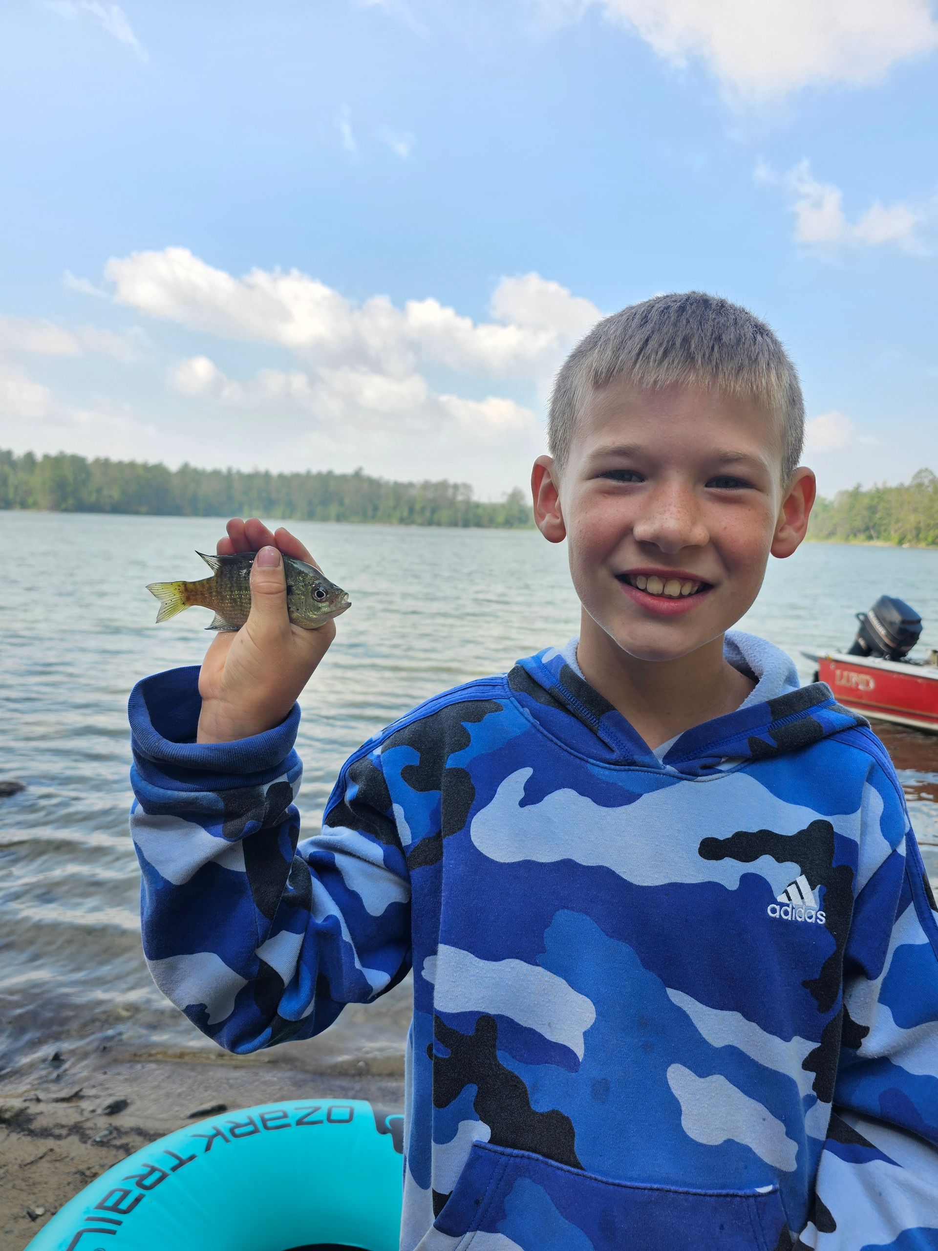 Smiling boy in blue camouflage shirt holding up a small fish by a lake with a kayak nearby