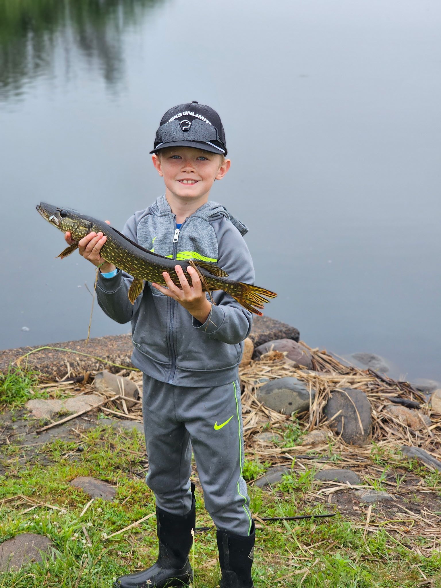 Child holding a large fish by a pond, wearing gray rain gear and boots, smiling at the camera
