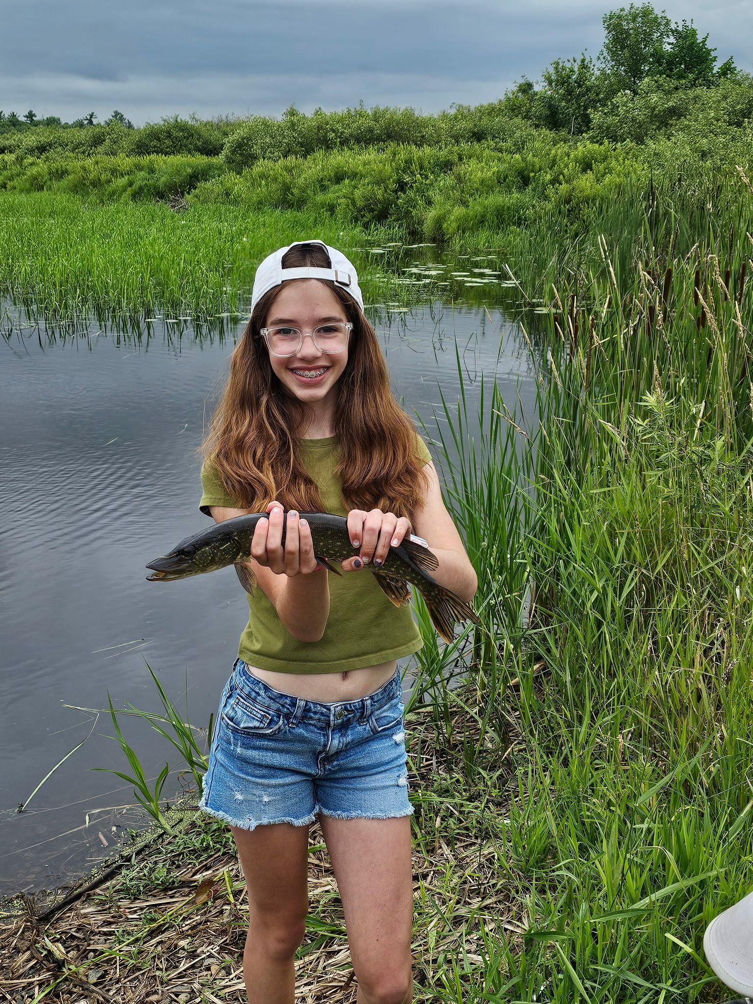 Woman holding a fish by a pond, wearing a green top, denim shorts, and a backward cap.
