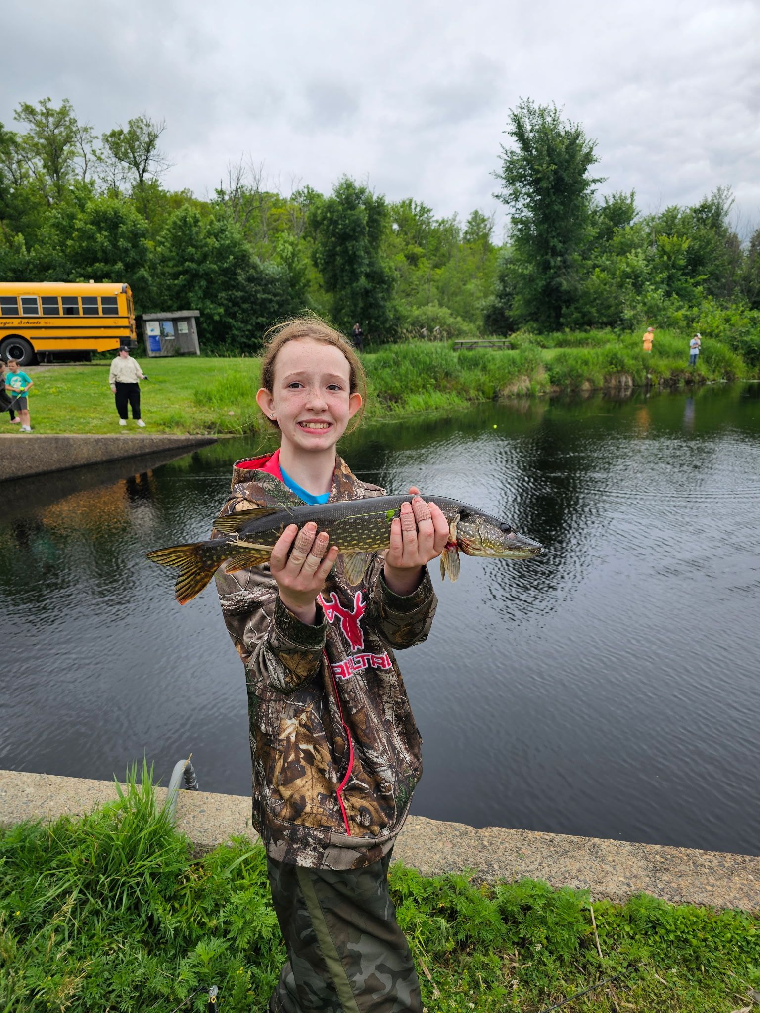 Person holding a fish by a pond, with a yellow bus and greenery in the background.