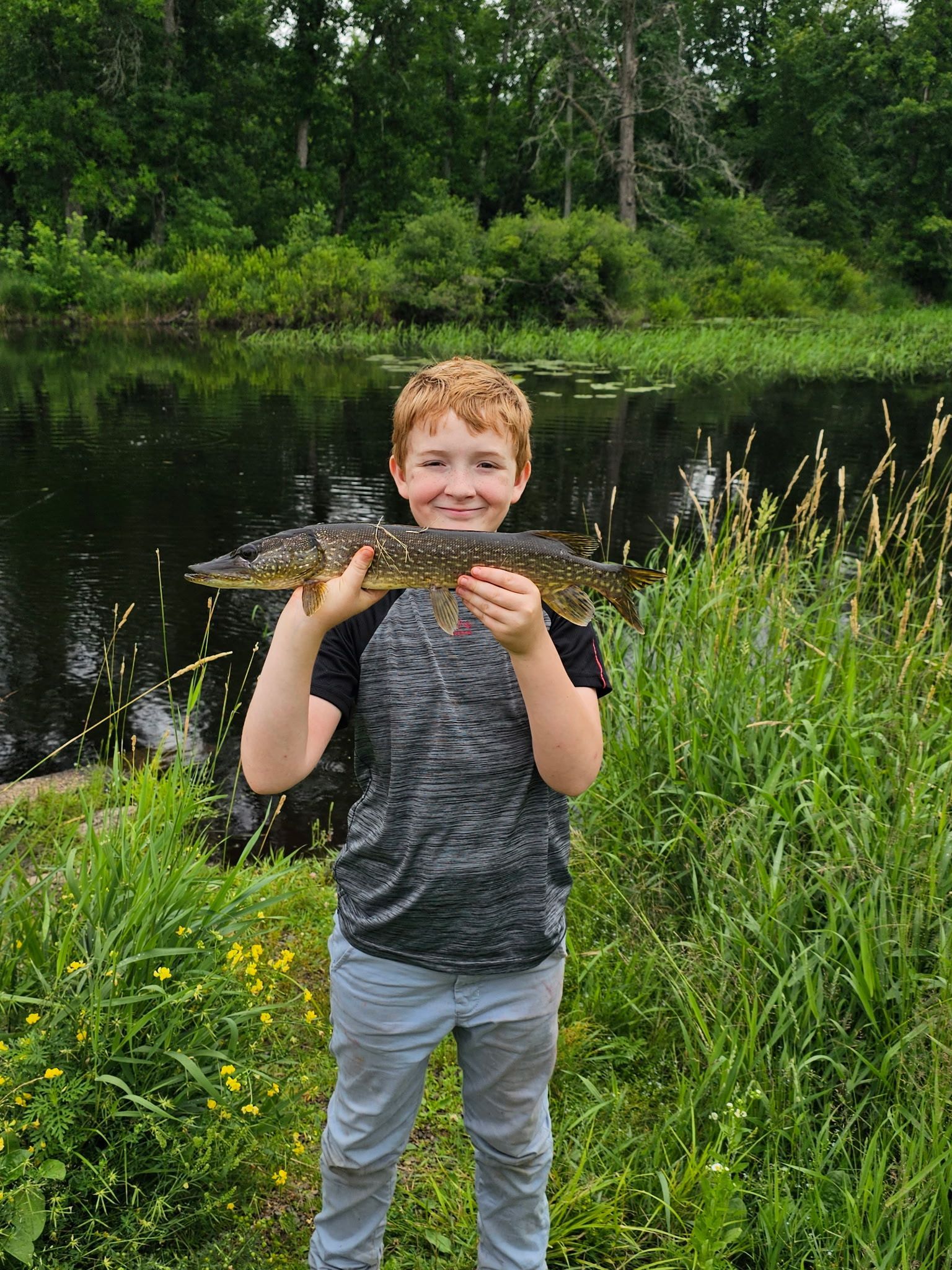 Child holding a fish by a pond, standing in tall grass with trees in the background.