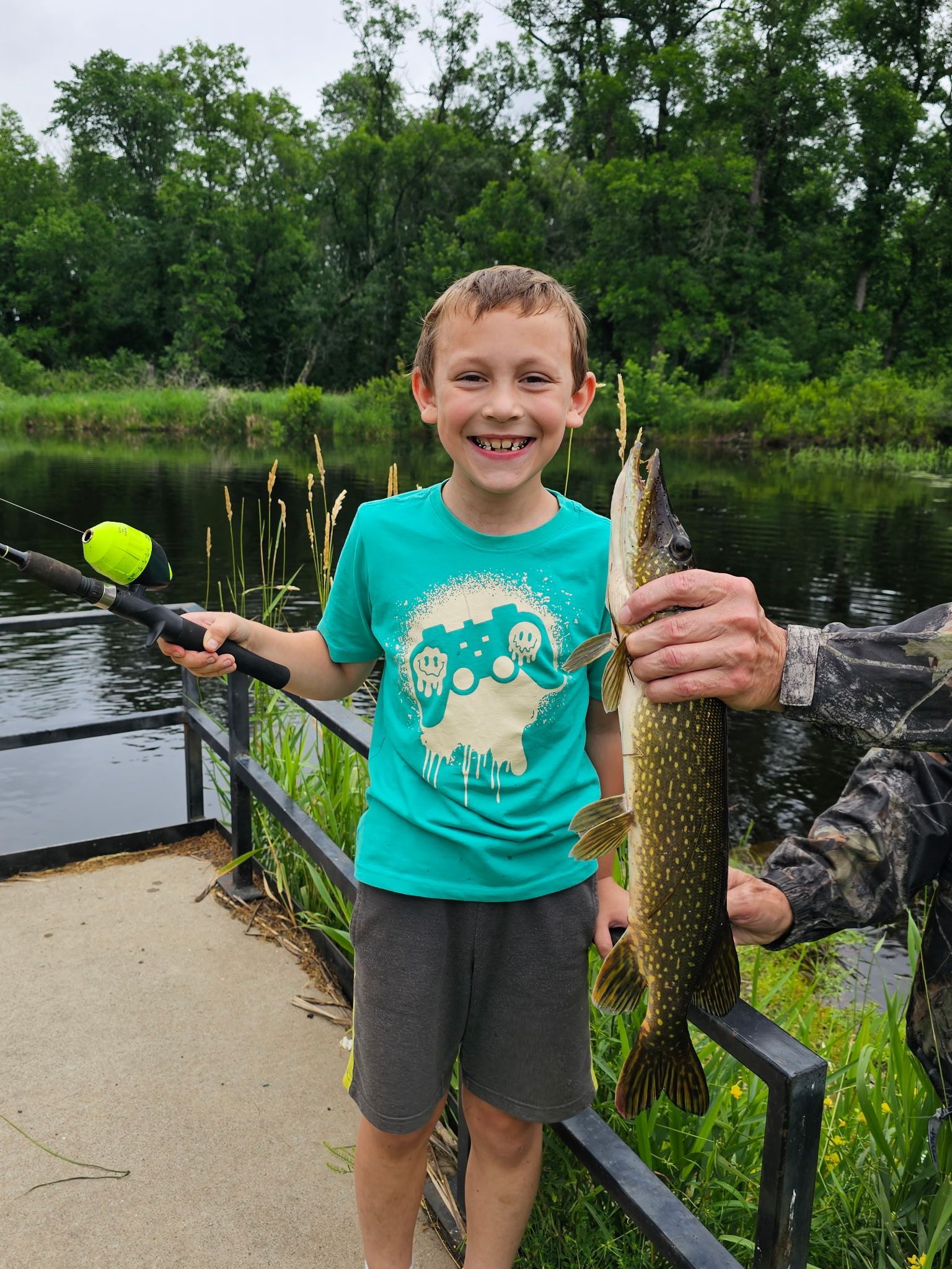 Smiling child holding a fishing rod and a fish beside a pond, with trees in the background.