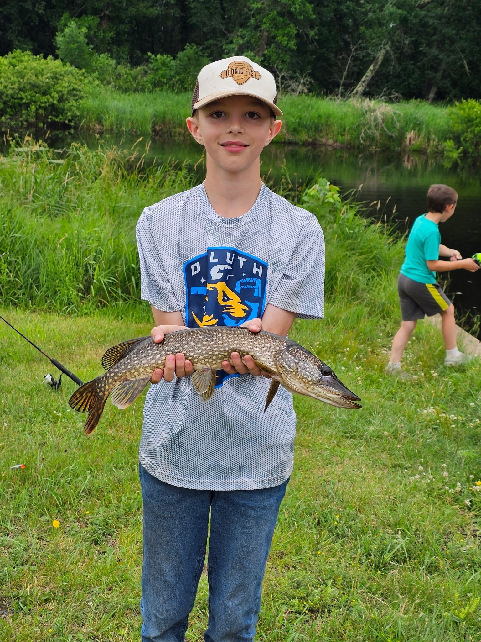 Boy holding a large fish in a grassy field, with another child fishing in the background.