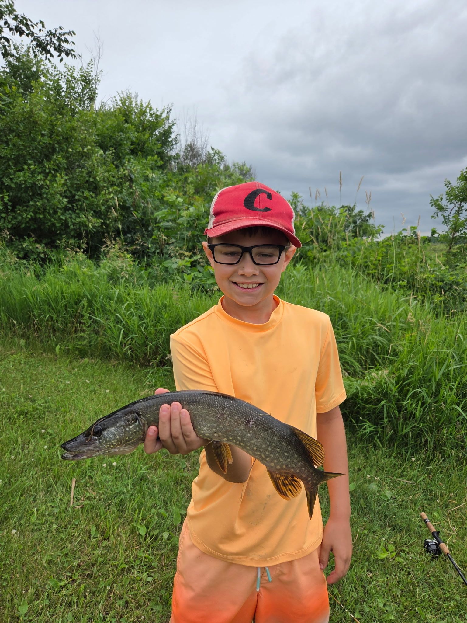 Person in orange shirt and red cap holding a fish outdoors on a grassy trail