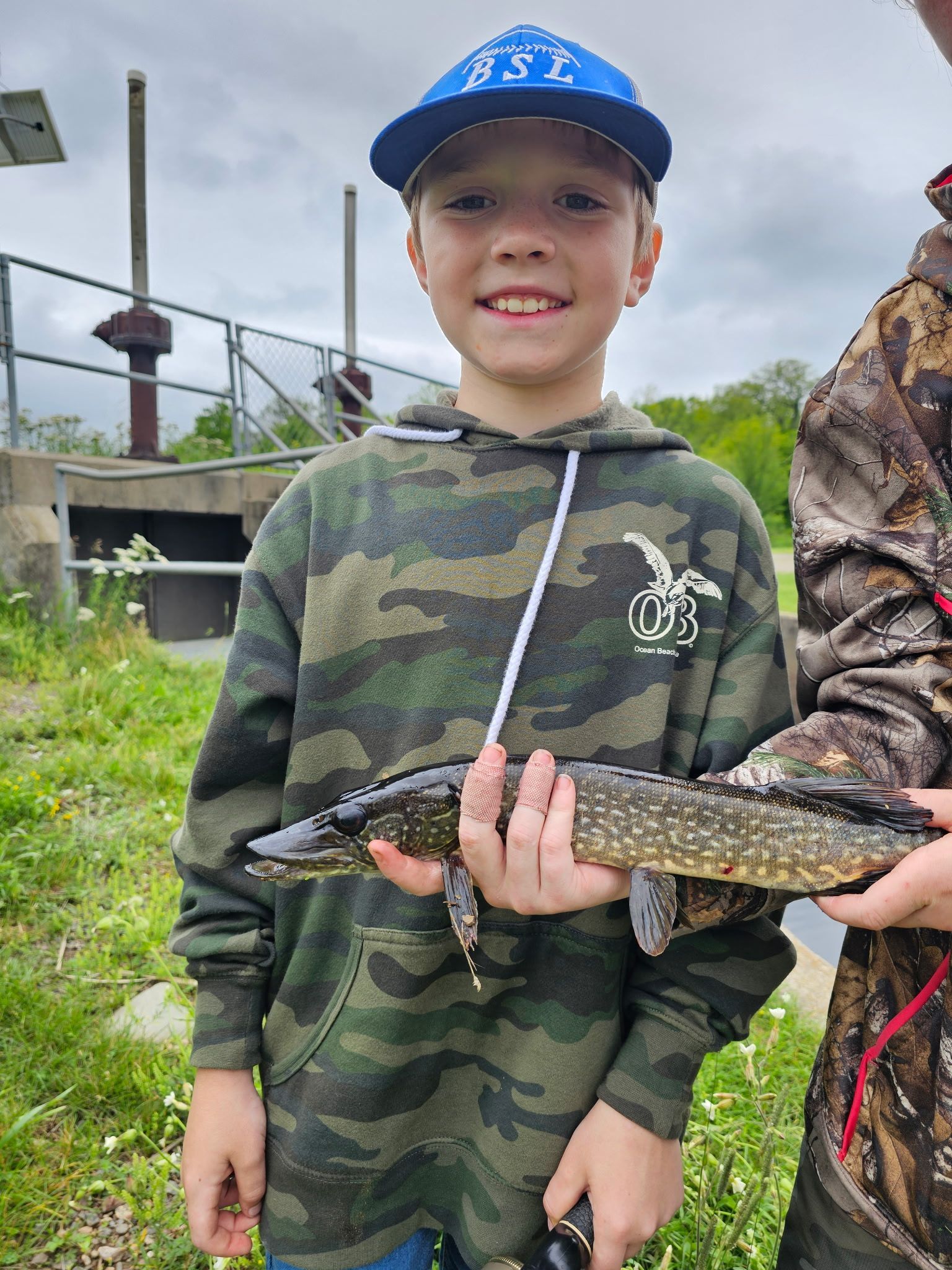 Child in blue cap and camo hoodie holding a fish outdoors by a dock